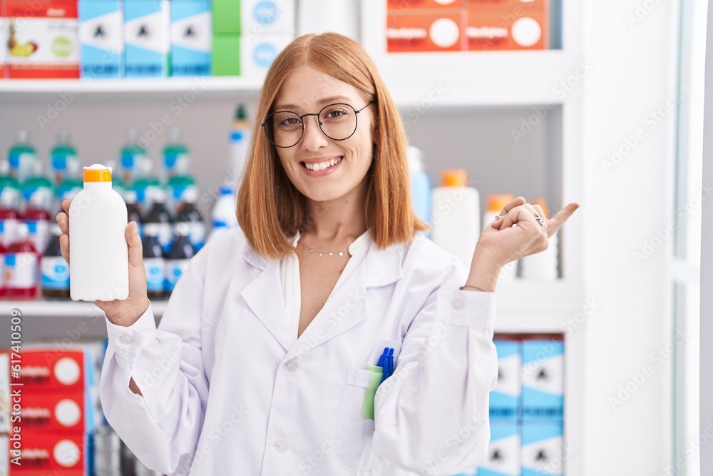 Young redhead woman working at pharmacy drugstore holding sun screen smiling happy pointing with hand and finger to the side