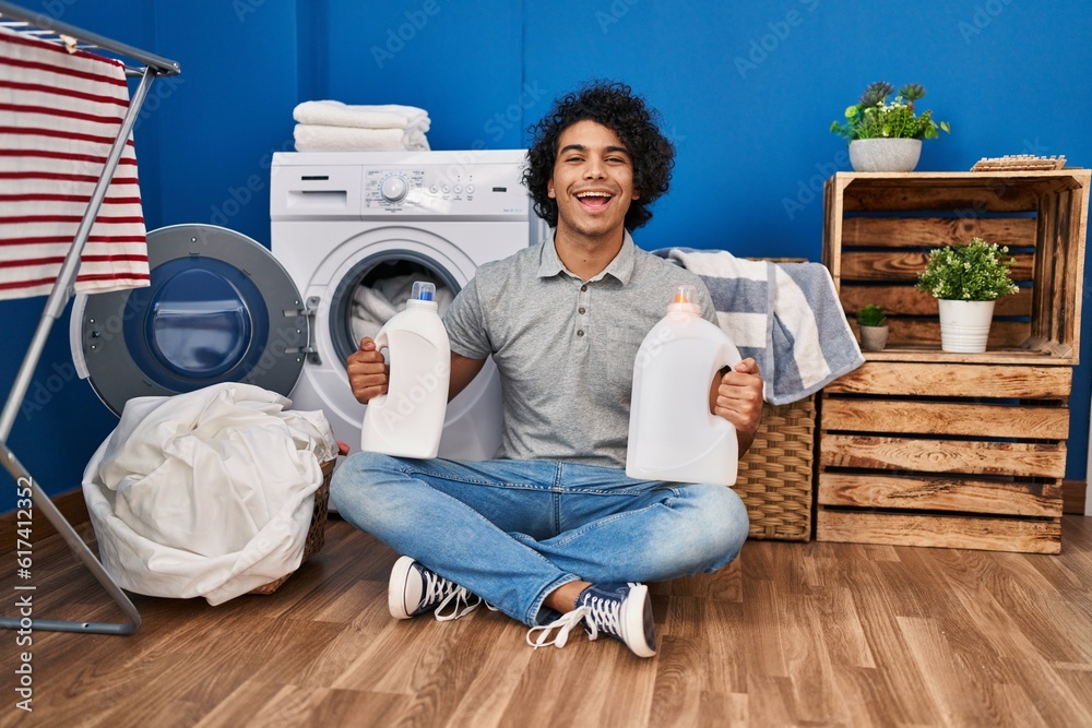 Hispanic man with curly hair doing laundry holding detergent bottles smiling and laughing hard out loud because funny crazy joke.
