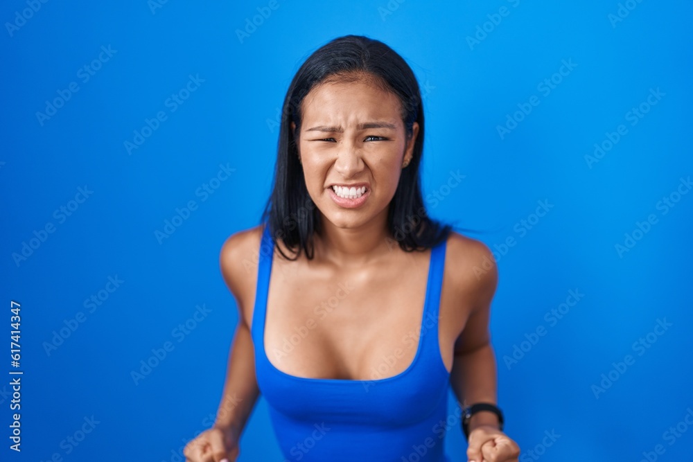 Hispanic woman standing over blue background angry and mad screaming frustrated and furious, shouting with anger. rage and aggressive concept. Hispanic woman standing over blue background angry and mad screaming frustrated and furious, shouting with anger. rage and aggressive concept.