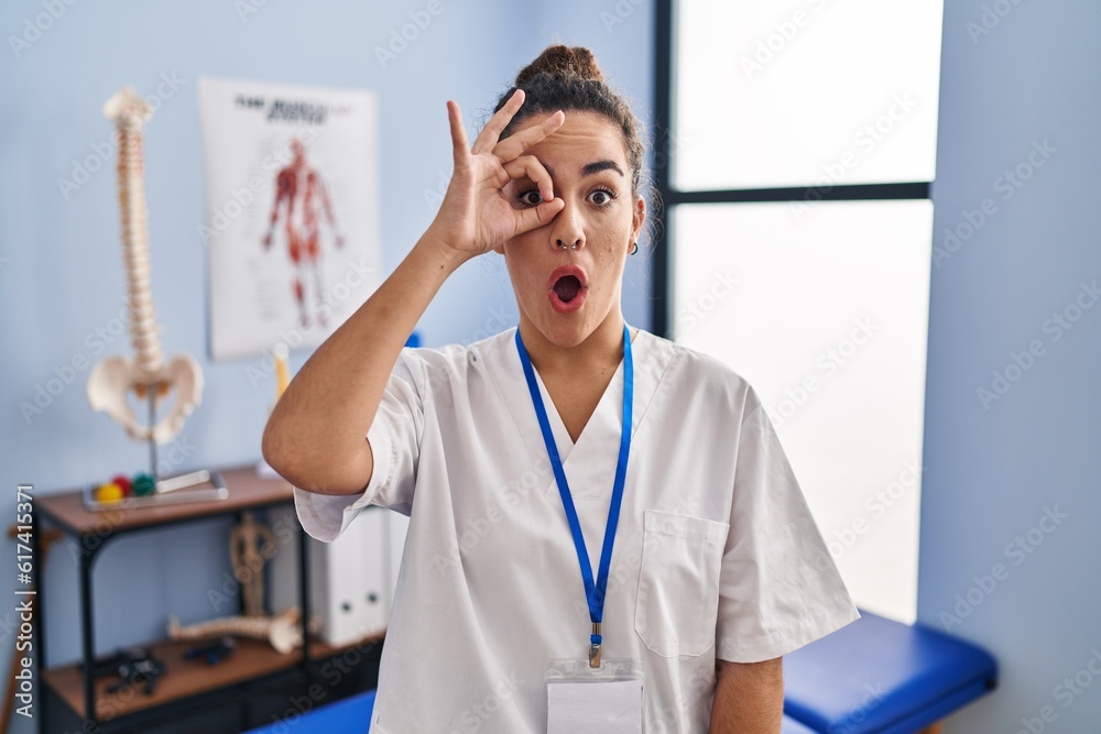 Young hispanic woman working at rehabilitation clinic doing ok gesture shocked with surprised face, eye looking through fingers. unbelieving expression. Young hispanic woman working at rehabilitation clinic doing ok gesture shocked with surprised face, eye looking through fingers. unbelieving expression.