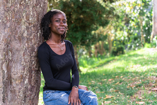 Young black woman with natural sister loc hair style sitting on a log in the park. Wearing blue jeans and a black top. No make up