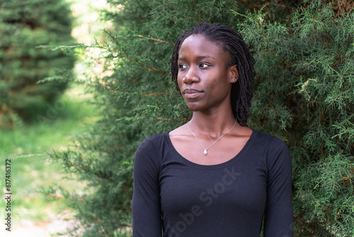 Young black woman with natural sister loc hairstyle smiling while standing in front of a pine tree at a park. No make up and wearing a black top