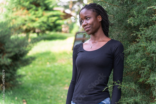 Young black woman with natural sister loc hairstyle smiling while standing in front of a pine tree at a park. No make up and wearing a black top