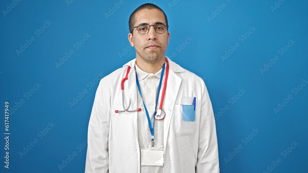 Fototapeta premium Young hispanic man doctor standing with serious expression over isolated blue background