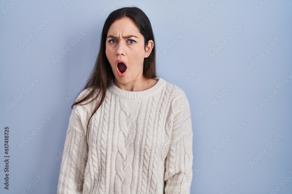 Young brunette woman standing over blue background in shock face, looking skeptical and sarcastic, surprised with open mouth Young brunette woman standing over blue background in shock face, looking skeptical and sarcastic, surprised with open mouth