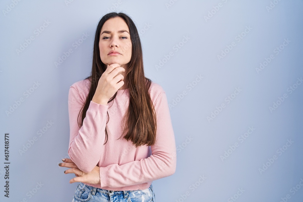 Young brunette woman standing over blue background looking confident at the camera with smile with crossed arms and hand raised on chin. thinking positive. Young brunette woman standing over blue background looking confident at the camera with smile with crossed arms and hand raised on chin. thinking positive.