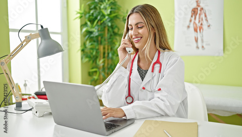 Young blonde woman doctor using laptop talking on telephone at clinic