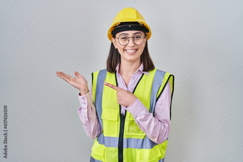 Hispanic girl wearing builder uniform and hardhat amazed and smiling to the camera while presenting with hand and pointing with finger. Hispanic girl wearing builder uniform and hardhat amazed and smiling to the camera while presenting with hand and pointing with finger.