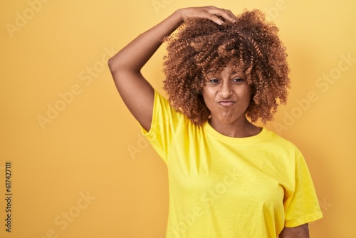 Young hispanic woman with curly hair standing over yellow background confuse and wonder about question. uncertain with doubt, thinking with hand on head. pensive concept.