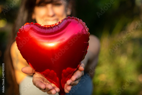 Mulher morena feliz e sorrindo segurando e oferecendo um coração vermelho em suas mãos, com ela e fundo desfocados. photo
