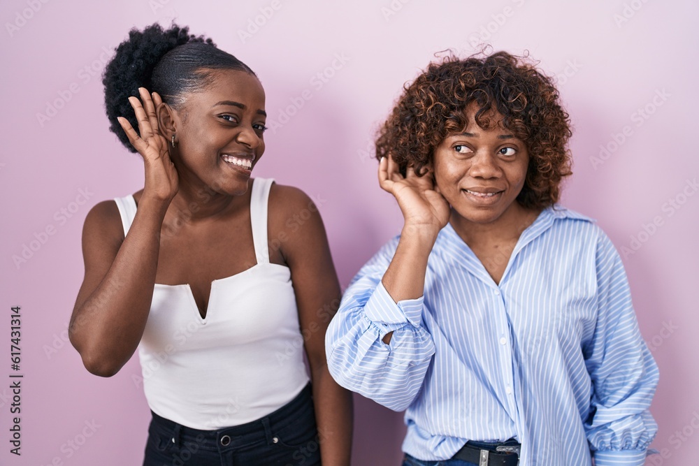 Two african women standing over pink background smiling with hand over ear listening an hearing to rumor or gossip. deafness concept. Two african women standing over pink background smiling with hand over ear listening an hearing to rumor or gossip. deafness concept.