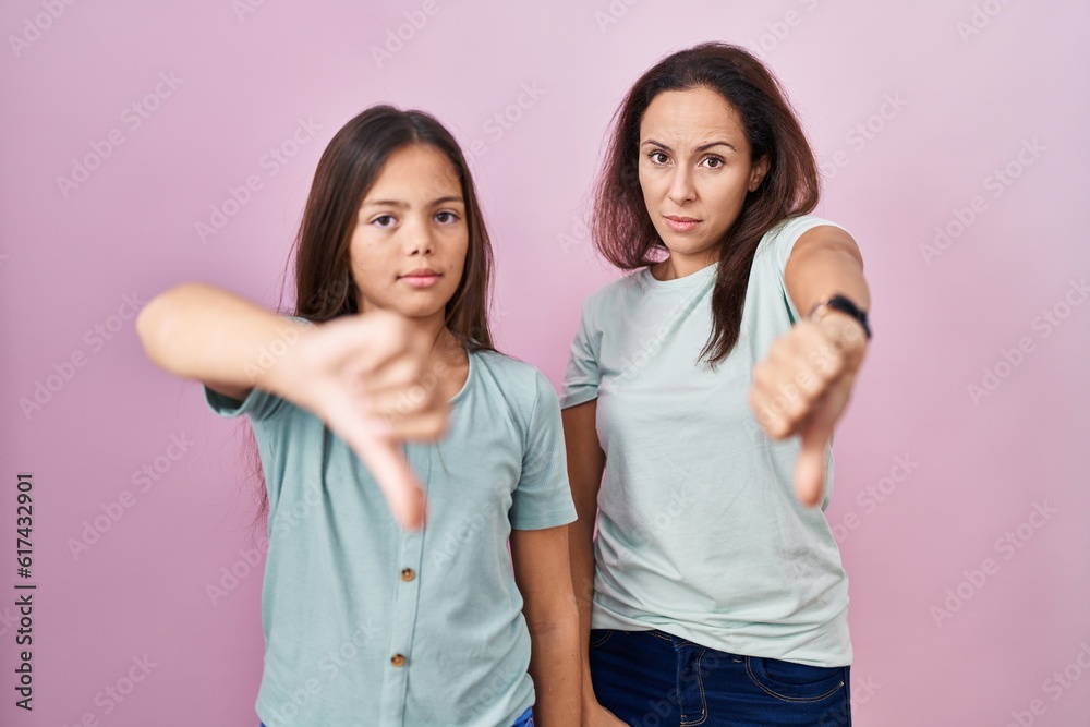 Young mother and daughter standing over pink background looking unhappy and angry showing rejection and negative with thumbs down gesture. bad expression. Young mother and daughter standing over pink background looking unhappy and angry showing rejection and negative with thumbs down gesture. bad expression.