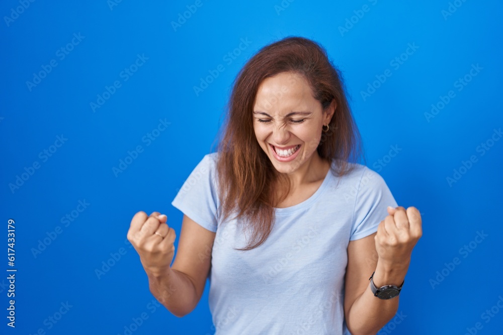 Fototapeta premium Brunette woman standing over blue background very happy and excited doing winner gesture with arms raised, smiling and screaming for success. celebration concept.