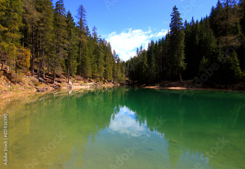 Landscape with the emerald smooth surface of Lago di Braies lake and pine trees on the shore against a blue sky with clouds in the Dolomites, Italy