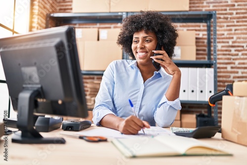 African american woman ecommerce business worker talking on smartphone writing on document at office