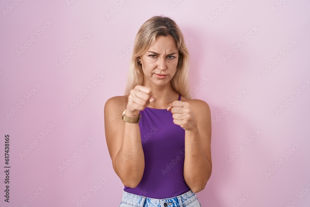 Young blonde woman standing over pink background ready to fight with fist defense gesture, angry and upset face, afraid of problem Young blonde woman standing over pink background ready to fight with fist defense gesture, angry and upset face, afraid of problem