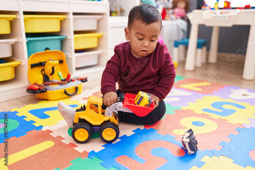 Adorable hispanic boy playing with tractor toy sitting on floor at kindergarten