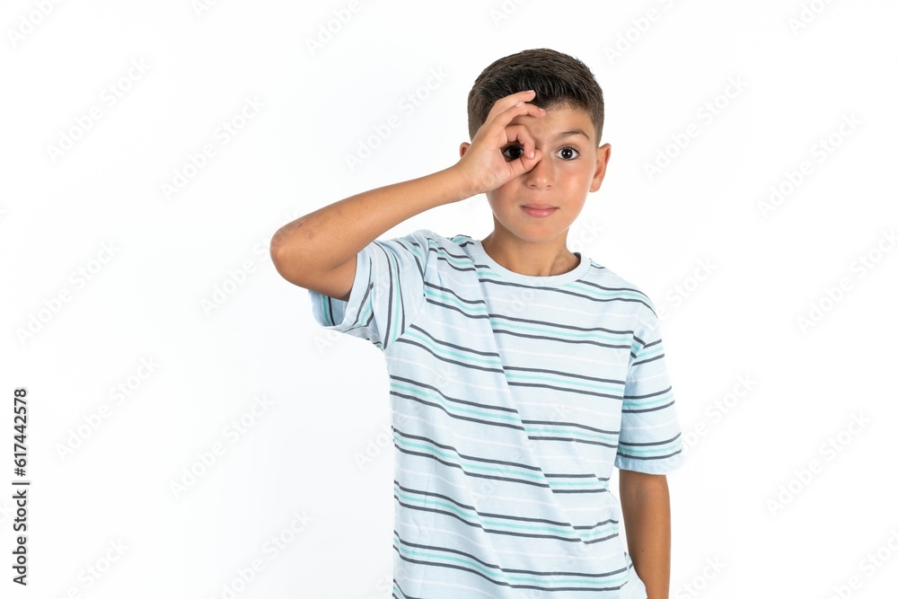 Little hispanic boy wearing striped T-shirt doing ok gesture with hand smiling, eye looking through fingers with happy face. Little hispanic boy wearing striped T-shirt doing ok gesture with hand smiling, eye looking through fingers with happy face.