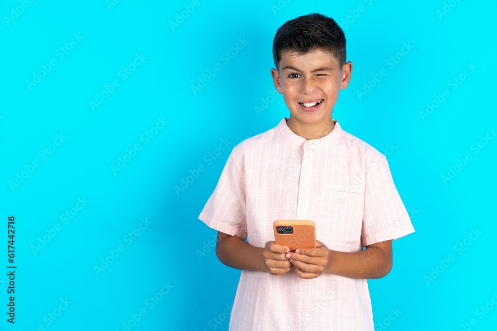 Excited Little hispanic boy wearing white shirt winking and eye hold smart phone use read social network news Excited Little hispanic boy wearing white shirt winking and eye hold smart phone use read social network news