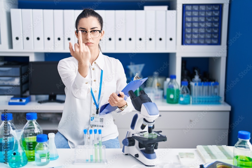 Young brunette woman working at scientist laboratory showing middle finger, impolite and rude fuck off expression