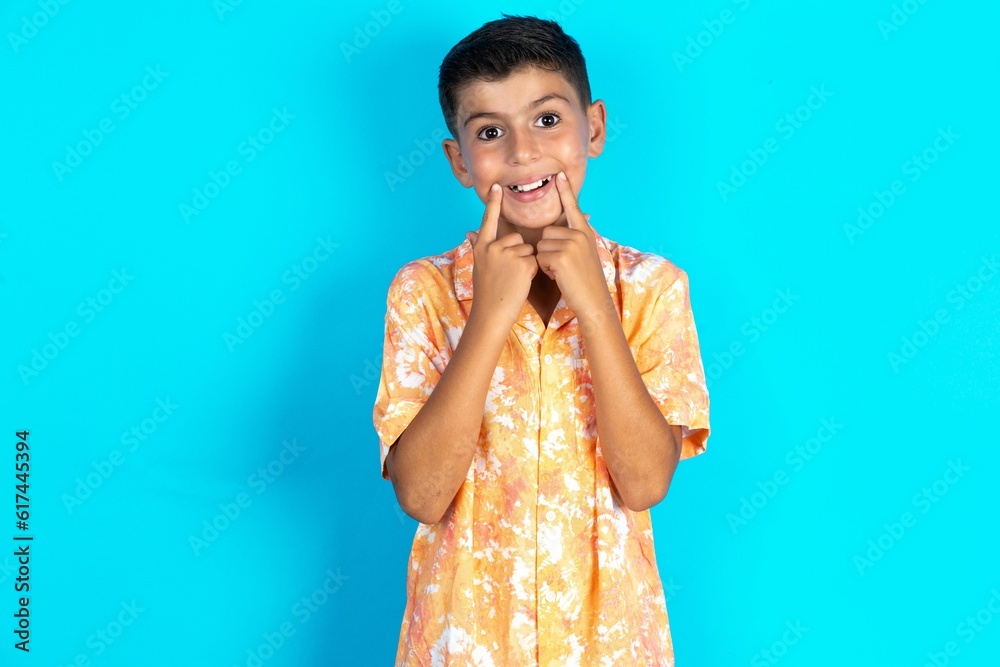 Happy Little hispanic kid boy wearing hawaiian shirt with toothy smile, keeps index fingers near mouth, fingers pointing and forcing cheerful smile Happy Little hispanic kid boy wearing hawaiian shirt with toothy smile, keeps index fingers near mouth, fingers pointing and forcing cheerful smile