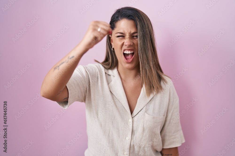 Blonde woman standing over pink background angry and mad raising fist frustrated and furious while shouting with anger. rage and aggressive concept. Blonde woman standing over pink background angry and mad raising fist frustrated and furious while shouting with anger. rage and aggressive concept.