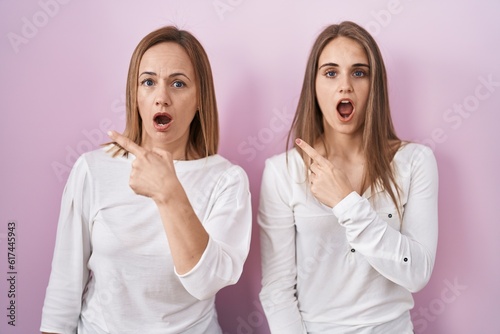Middle age mother and young daughter standing over pink background surprised pointing with finger to the side, open mouth amazed expression.