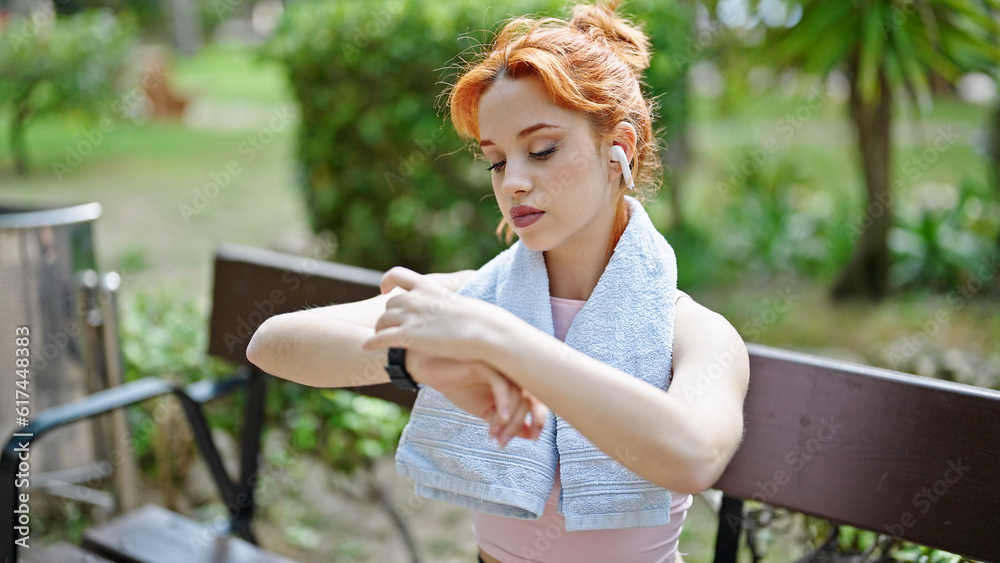 Fototapeta premium Young redhead woman using smartwatch and earphones sitting on bench at park
