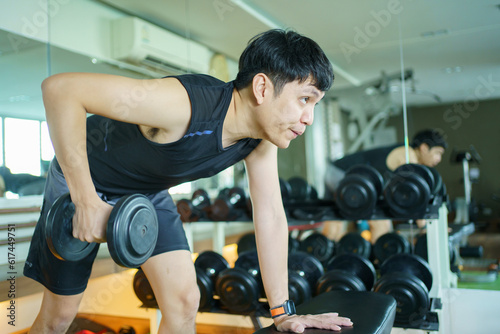 Happy cheerful Asian sportsman doing a weight training exercise at triceps muscular by weight lifting. Asian sporty man doing a dumbbell weight lifting workout in an indoor fitness gym.