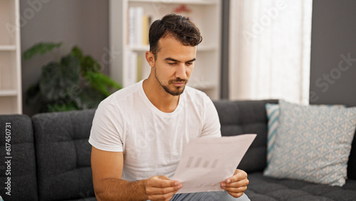 Young hispanic man reading bill sitting on sofa at home