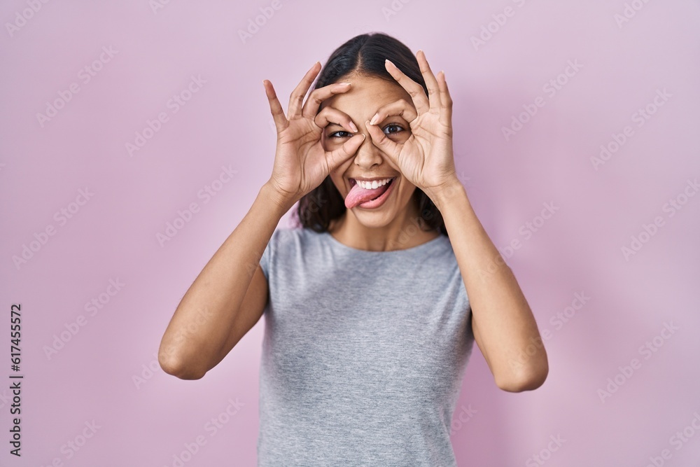 Young brazilian woman wearing casual t shirt over pink background doing ok gesture like binoculars sticking tongue out, eyes looking through fingers. crazy expression. Young brazilian woman wearing casual t shirt over pink background doing ok gesture like binoculars sticking tongue out, eyes looking through fingers. crazy expression.