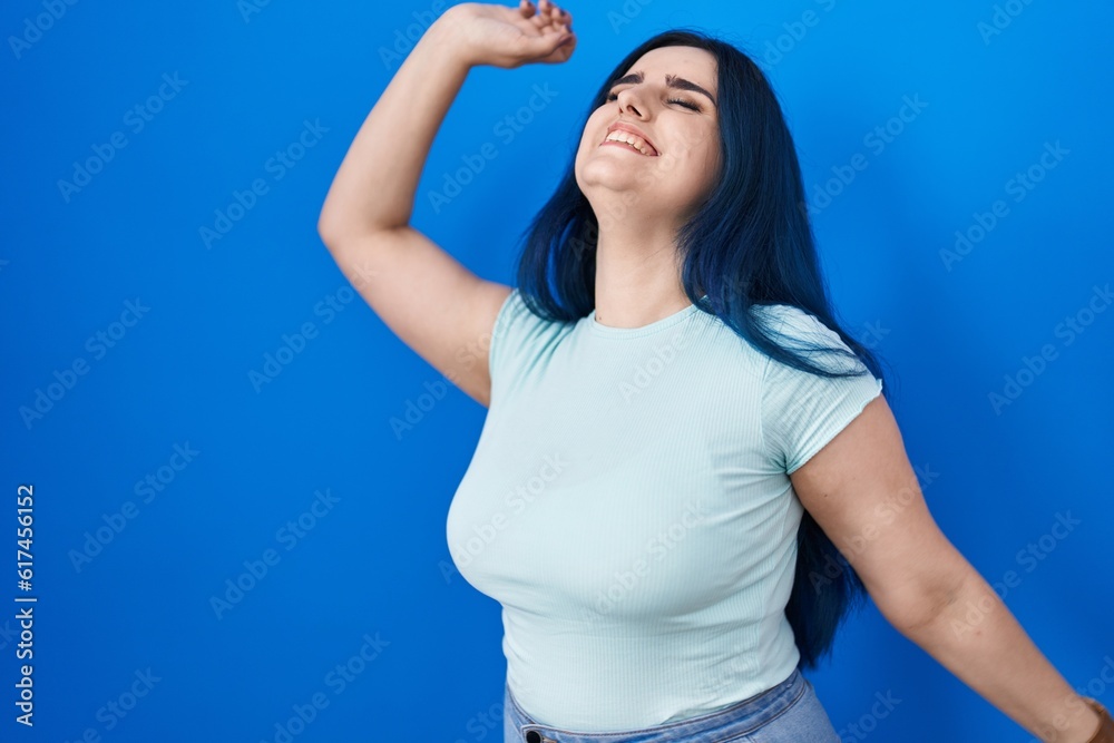 Young modern girl with blue hair standing over blue background dancing happy and cheerful, smiling moving casual and confident listening to music Young modern girl with blue hair standing over blue background dancing happy and cheerful, smiling moving casual and confident listening to music