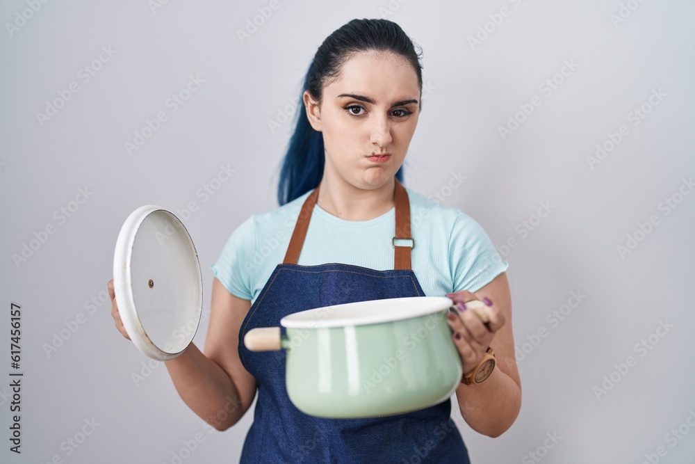 Young modern girl with blue hair wearing apron holding cooking pot skeptic and nervous, frowning upset because of problem. negative person. Young modern girl with blue hair wearing apron holding cooking pot skeptic and nervous, frowning upset because of problem. negative person.