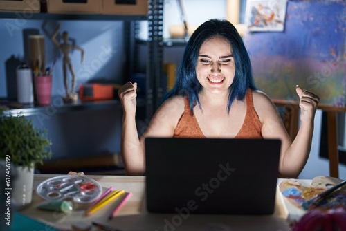 Young modern girl with blue hair sitting at art studio with laptop at night very happy and excited doing winner gesture with arms raised, smiling and screaming for success. celebration concept.