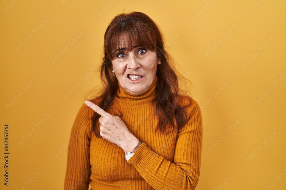 Middle age hispanic woman standing over yellow background pointing aside worried and nervous with forefinger, concerned and surprised expression Middle age hispanic woman standing over yellow background pointing aside worried and nervous with forefinger, concerned and surprised expression