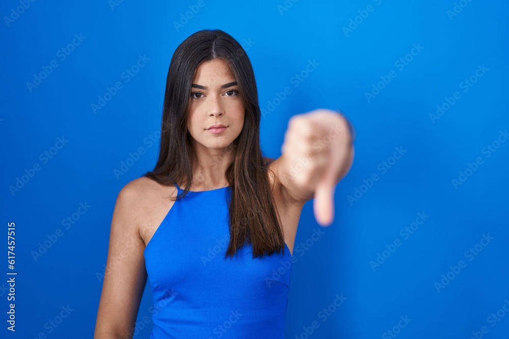 Hispanic woman standing over blue background looking unhappy and angry showing rejection and negative with thumbs down gesture. bad expression.