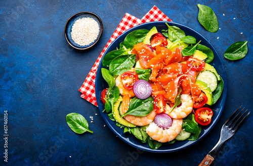Fresh salad for keto diet with salmon, avocado, spinach, cucumber, shrimps, tomato, cashew, sesame seeds. Low-carbohydrate lunch rich in healthy fats. Blue stone table background, top view