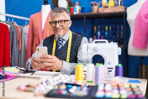 Middle age grey-haired man tailor smiling confident using smartphone at clothing factory