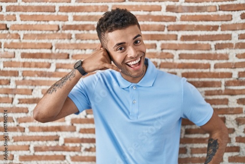 Brazilian young man standing over brick wall smiling doing phone gesture with hand and fingers like talking on the telephone. communicating concepts.