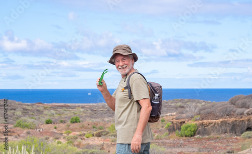 Happy active senior man in hat and sunglasses hiking in countryside close to the sea enjoying healthy lifestyle and freedom
