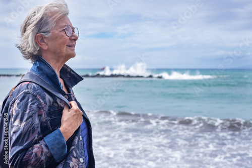 Portrait of senior gray haired woman in denim jacket and backpack standing in front to the sea looking at horizon enjoying relax and retirement #617461945