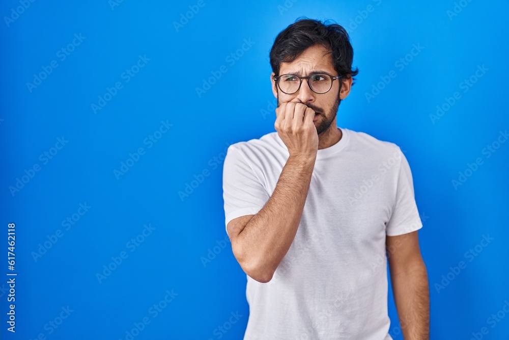 Handsome latin man standing over blue background looking stressed and nervous with hands on mouth biting nails. anxiety problem. Handsome latin man standing over blue background looking stressed and nervous with hands on mouth biting nails. anxiety problem.