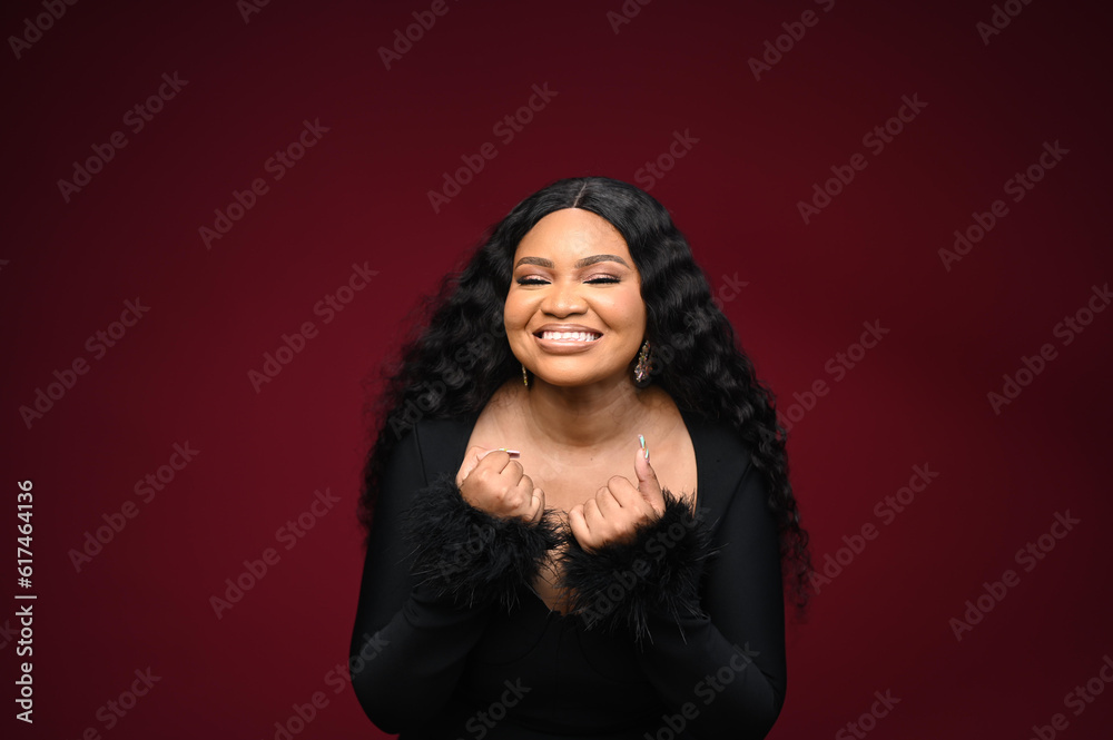 Amused happy grateful charming African-American woman in evening black dress fist chest position thankful smiling broadly appreciating awesome touching heartwarming gift, standing red background