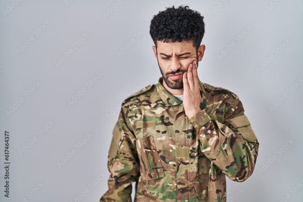 Arab man wearing camouflage army uniform touching mouth with hand with painful expression because of toothache or dental illness on teeth. dentist