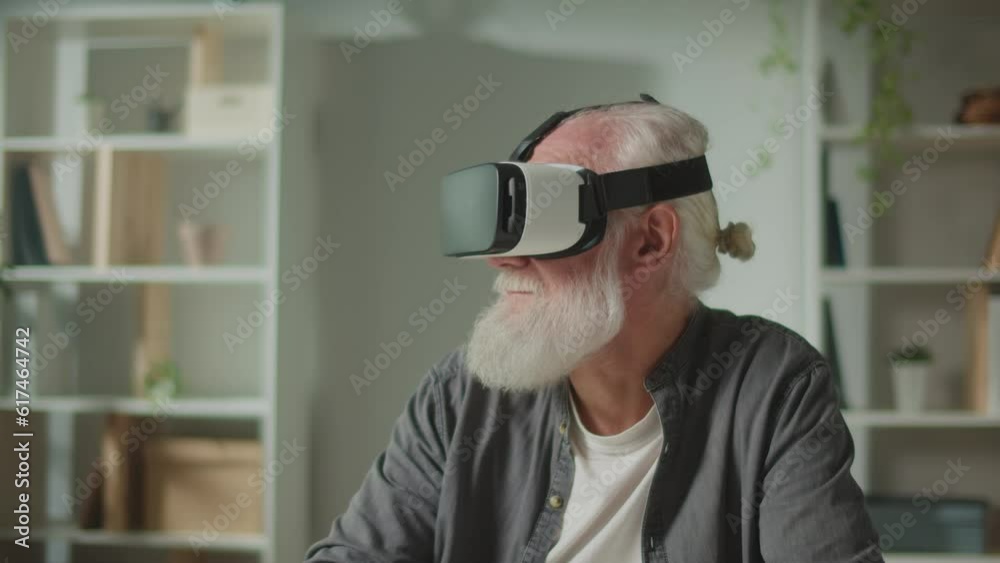 A Modern Old Man With Gray Beard in VR Glasses Examines Everything Around. An Elderly Man in VR Glasses Sitting at a Table at Home.VR to Help the Elderly. A Modern Old Man With Gray Beard in VR Glasses Examines Everything Around. An Elderly Man in VR Glasses Sitting at a Table at Home.VR to Help the Elderly.