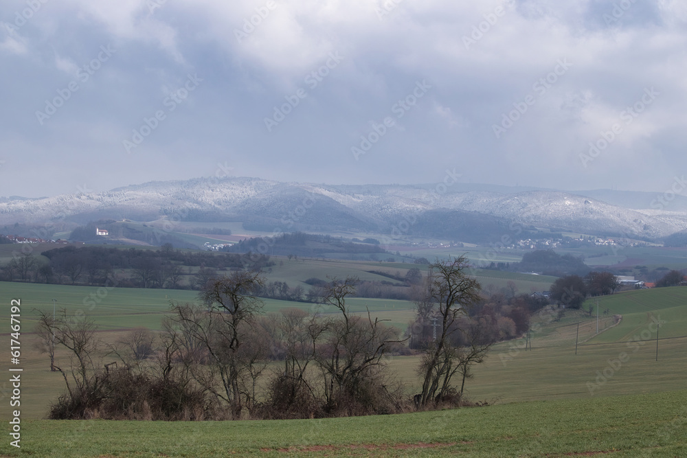 Obraz premium Trees in a green field with sun shining on a snow covered hill in the background on a cloudy winter day in Rhineland Palatinate, germany.