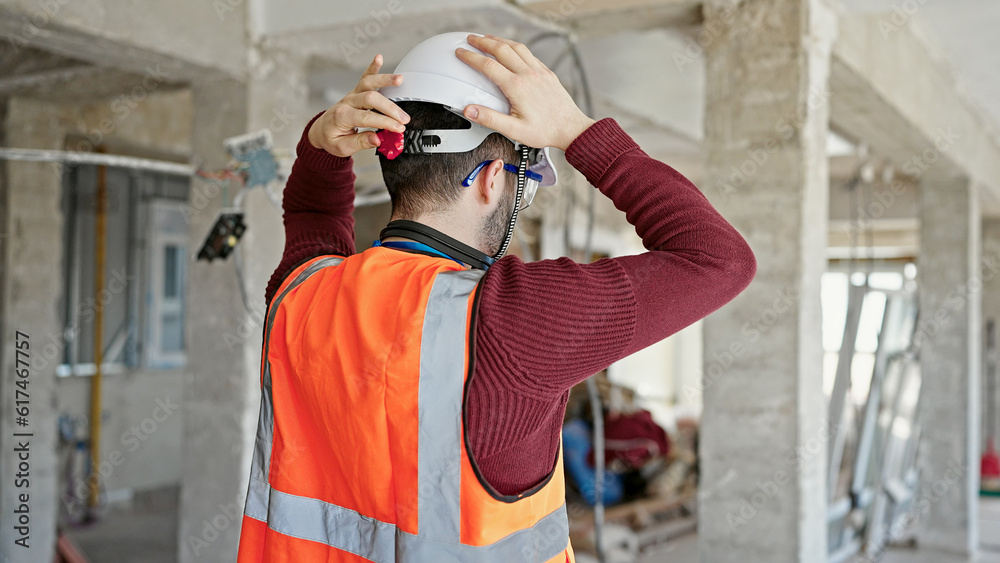 Fototapeta premium Young hispanic man builder wearing hardhat standing backwards at construction site
