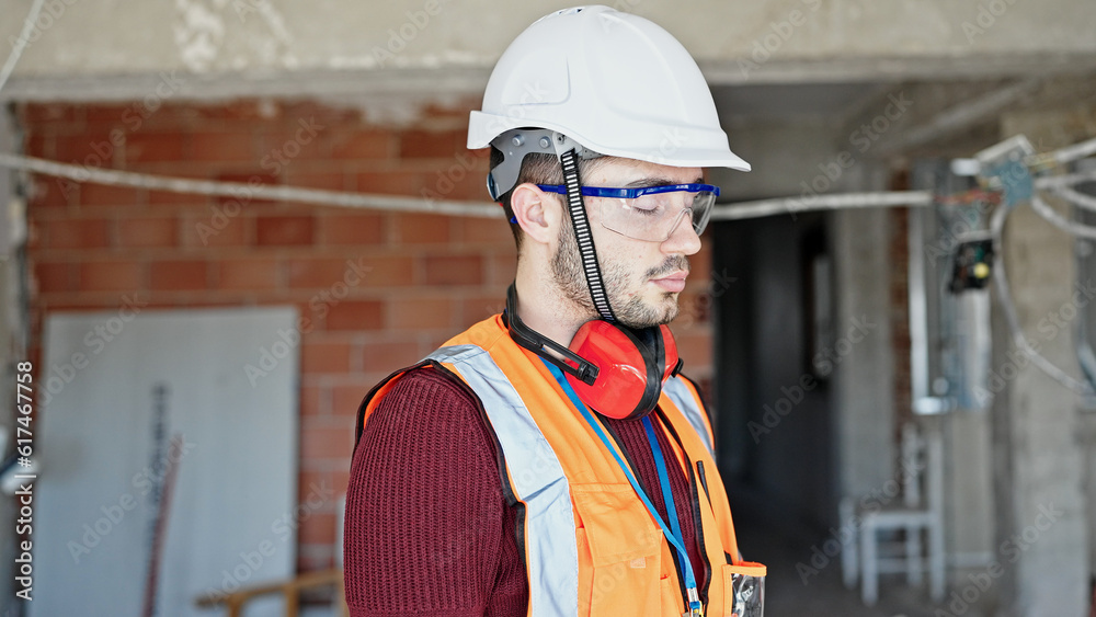 Young hispanic man builder standing with serious face at construction site