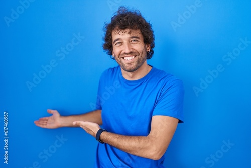 Photography Hispanic young man standing over blue background inviting to enter smiling natur