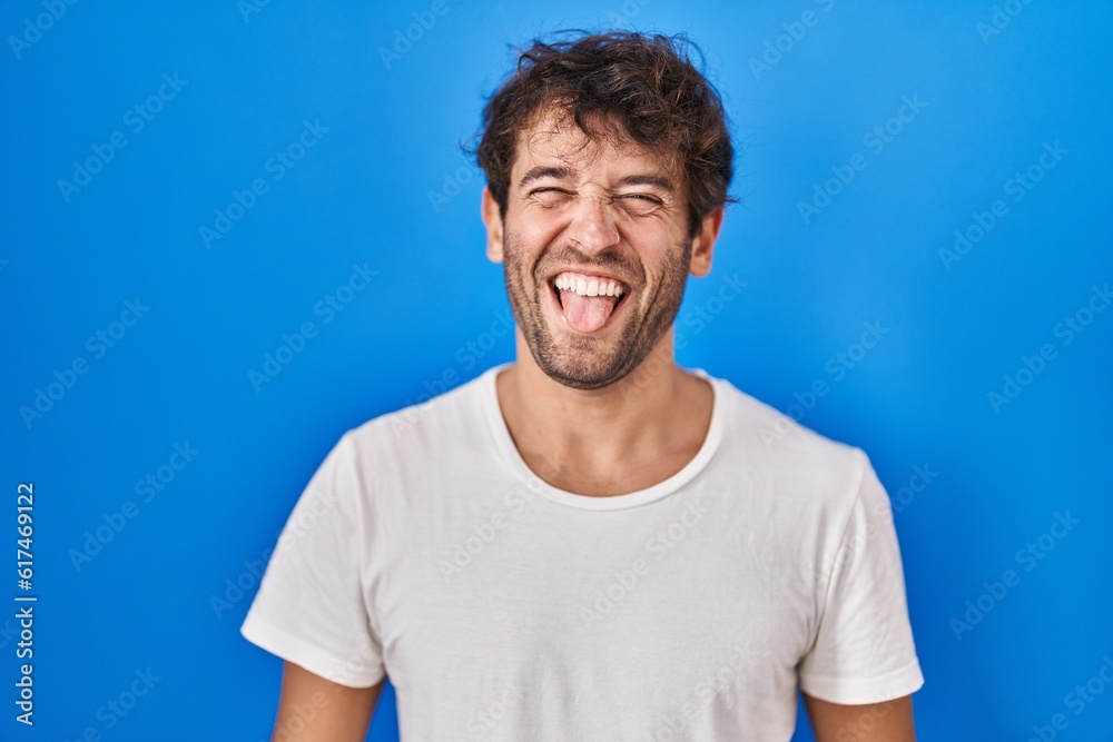 Fototapeta premium Hispanic young man standing over blue background sticking tongue out happy with funny expression. emotion concept.